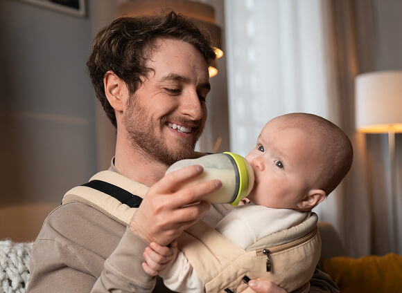 Man feeding a baby from a bottle while holding the baby in a carrier.